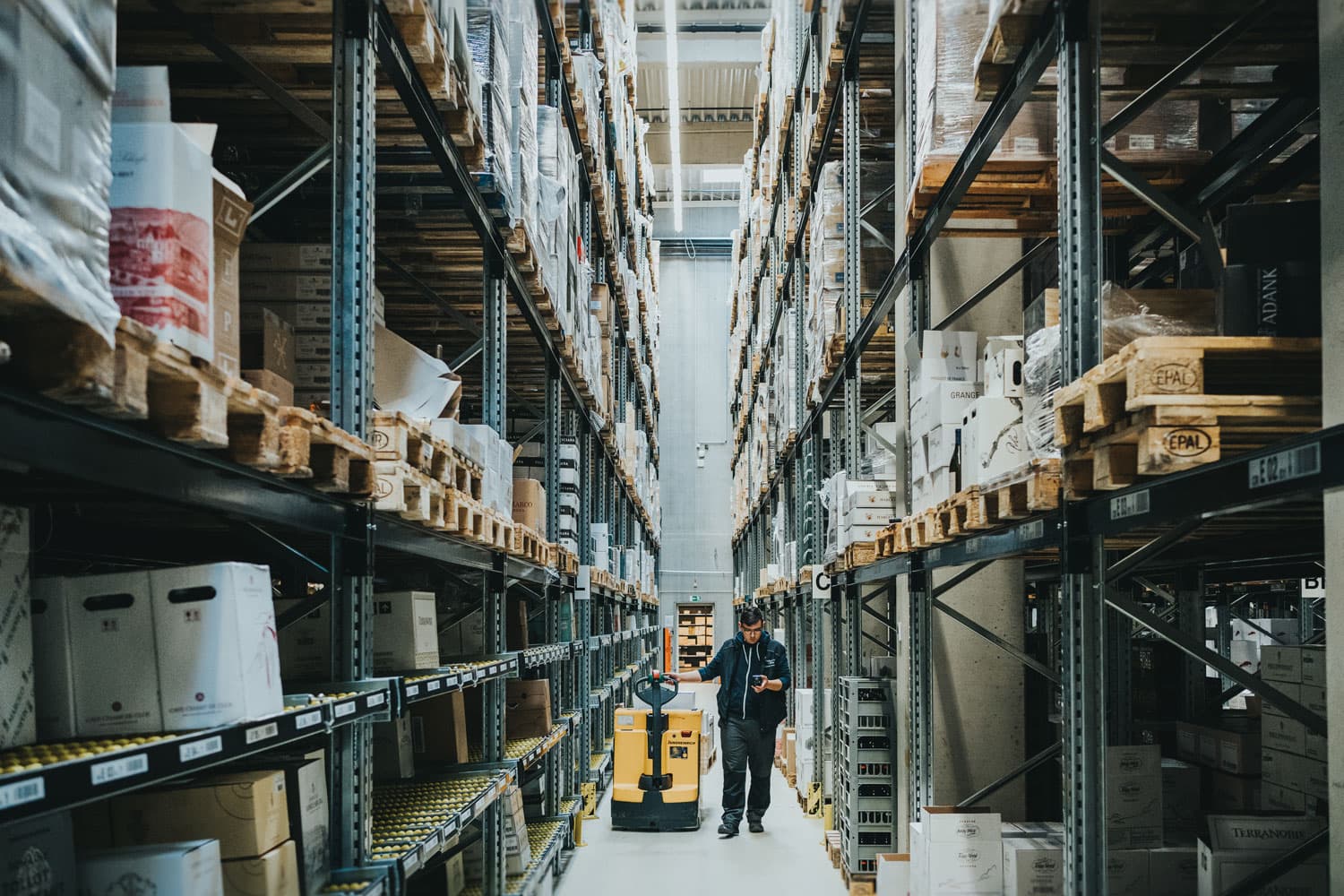 Lagermitarbeiter%2C%20der%20einen%20gelben%20Hubwagen%20f%E4hrt%20und%20ein%20Handger%E4t%20%FCberpr%FCft%2C%20umgeben%20von%20hohen%20Regalen%20mit%20Paletten%20und%20Lagerkartons