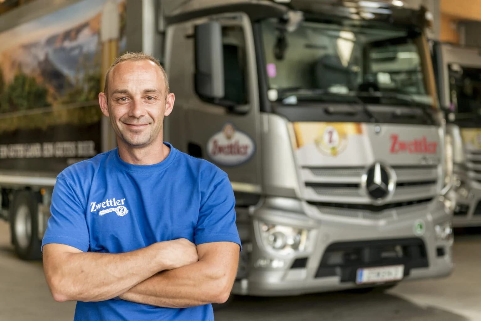 Lkw-Fahrer%20im%20blauen%20Zwettler-Shirt%20steht%20mit%20verschr%E4nkten%20Armen%20vor%20einem%20silbernen%20Lieferfahrzeug%20in%20einer%20Logistikhalle.