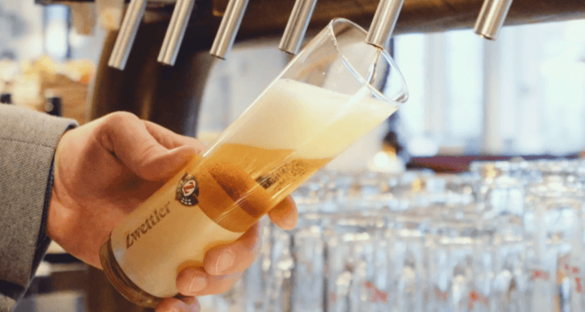 Hand%20zapft%20ein%20frisch%20gezapftes%20Bier%20mit%20Schaumkrone%20aus%20mehreren%20Zapfh%E4hnen%20in%20ein%20Markenbierglas%20an%20der%20Theke