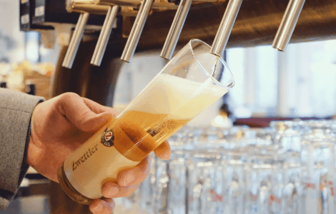 Hand%20zapft%20ein%20frisch%20gezapftes%20Bier%20mit%20Schaumkrone%20aus%20mehreren%20Zapfh%E4hnen%20in%20ein%20Markenbierglas%20an%20der%20Theke