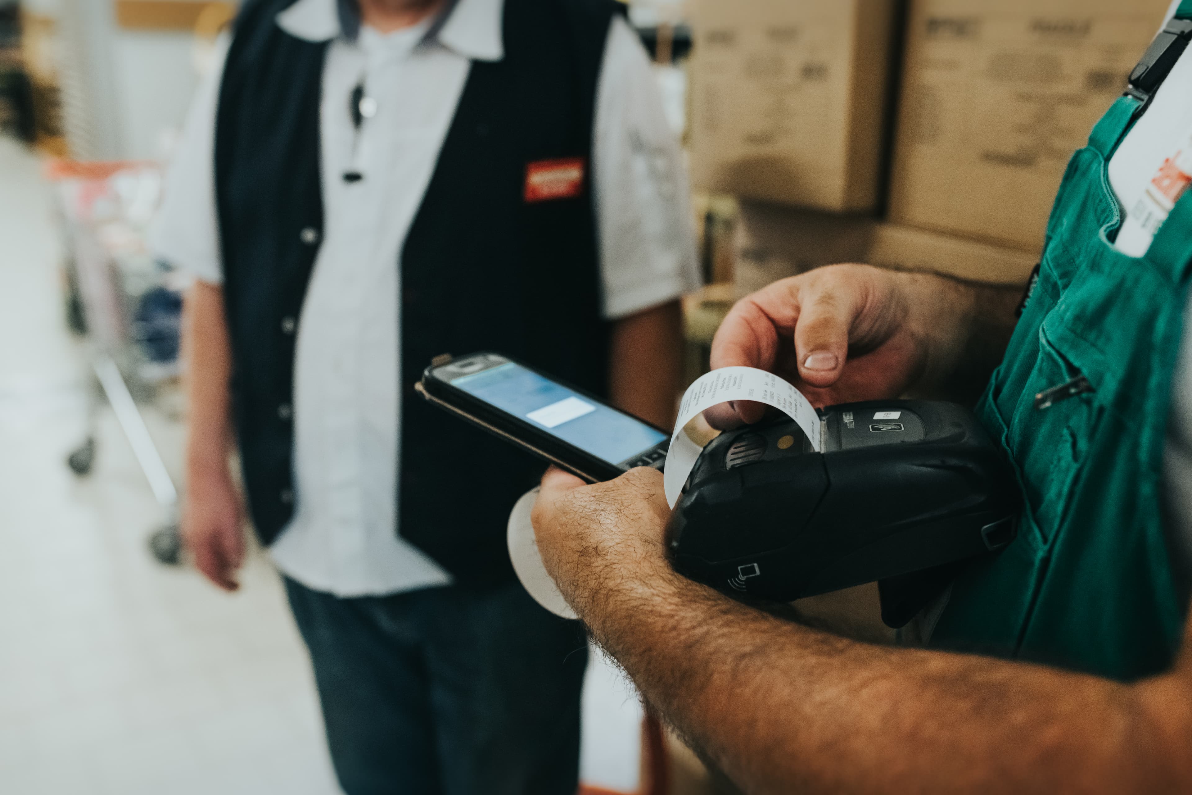 Nahaufnahme%20von%20zwei%20Personen%20in%20einem%20Lager%20oder%20Gesch%E4ftsumfeld.%20Eine%20Person%20h%E4lt%20ein%20mobiles%20Ger%E4t%20und%20einen%20kleinen%20Drucker%2C%20aus%20dem%20ein%20Beleg%20kommt.%20Im%20Hintergrund%20steht%20eine%20weitere%20Person%20vor%20gestapelten%20Kartons.